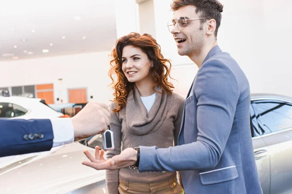 Ausgeschnittene Ansicht eines Autohändlers mit Schlüsseln in der Nähe eines glücklichen Mannes mit Brille und lockiger Frau — Stockfoto