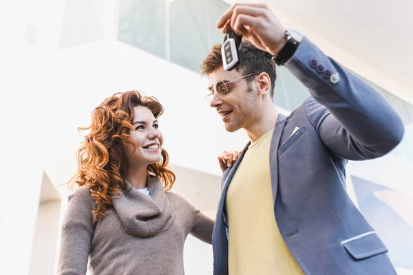 Bottom view of cheerful man in glasses looking at girl while holding keys in car showroom — Stock Photo