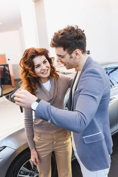 Happy man in glasses standing with cheerful curly woman and holding keys in car showroom — Stock Photo