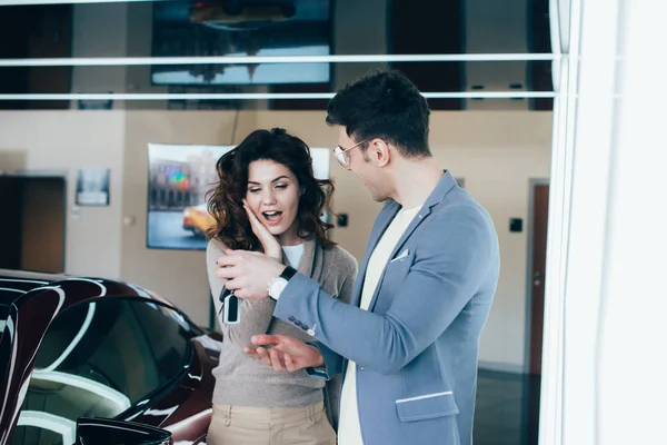 Cheerful man holding keys near surprised curly girl while standing near red automobile — Stock Photo