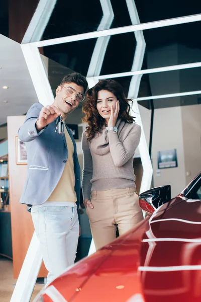 Alegre hombre sosteniendo llaves mientras de pie con sorprendido rizado chica cerca de coche - foto de stock