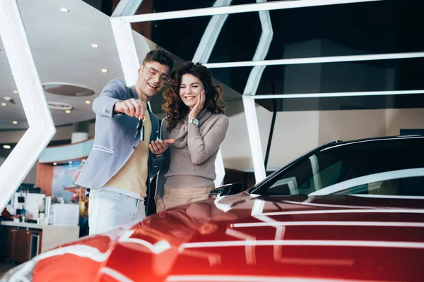 Веселий чоловік в окулярах тримає ключі, стоячи з усміхненою дівчиною біля червоного автомобіля — стокове фото