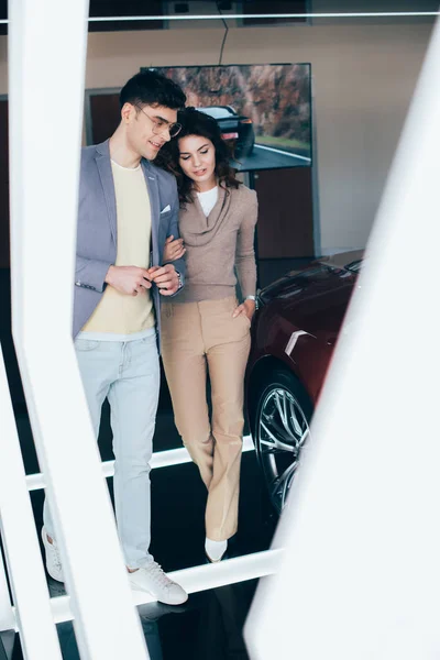 Selective focus of stylish man in glasses walking with curly girl near red automobile — Stock Photo