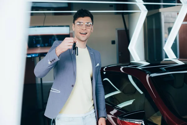 Веселий чоловік в окулярах тримає ключі, стоячи біля червоного автомобіля — стокове фото