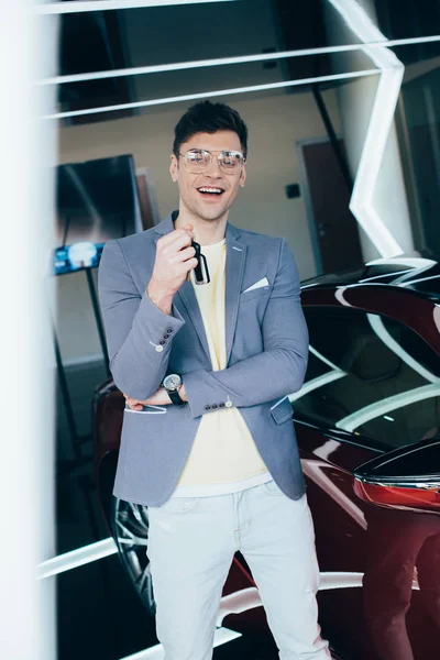 Selective focus of happy stylish man in glasses holding keys while standing near red automobile — Stock Photo