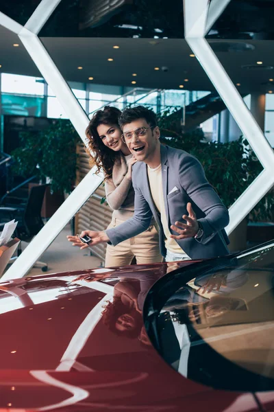 Hombre emocionado sonriendo con chica rizada feliz cerca de coche rojo - foto de stock