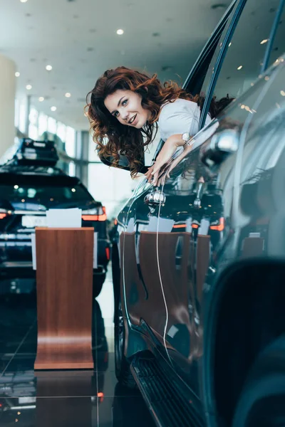 Selective focus of cheerful curly woman smiling while looking out of window in automobile — Stock Photo