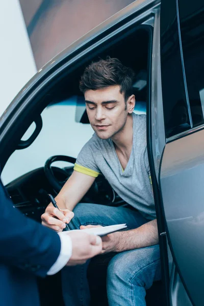 Recortado vista de concesionario de coches sujetando portapapeles mientras hombre guapo firma contrato - foto de stock