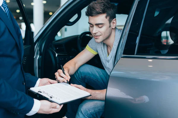 Abgeschnittene Ansicht von Autohändler hält Klemmbrett, während fröhlicher Mann Vertrag unterschreibt — Stockfoto