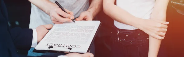 Panoramic shot of car dealer holding clipboard while man signing contract near woman — Stock Photo