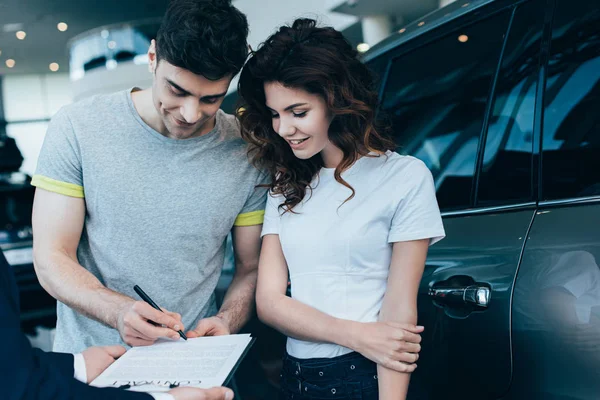 Обрезанный вид автомобиля дилер держит планшет в то время как счастливый человек подписывает контракт рядом с привлекательной кудрявой женщиной — стоковое фото
