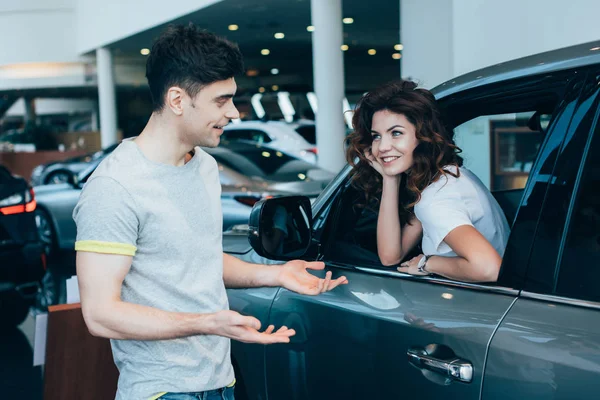 Cheerful woman sitting in automobile near handsome man standing and gesturing in car showroom — Stock Photo
