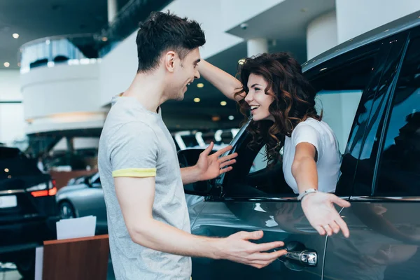 Cheerful woman sitting in automobile with outstretched hands near handsome man — Stock Photo
