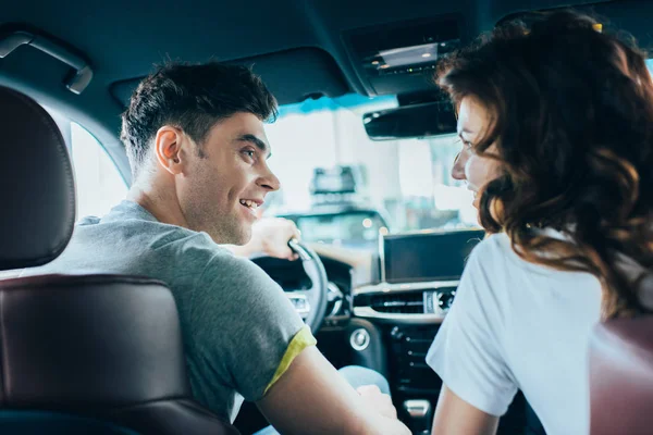 Selective focus of handsome man looking at curly cheerful woman while sitting in automobile — Stock Photo
