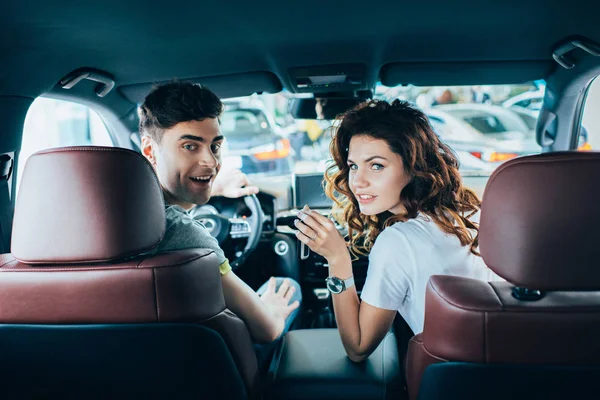 Selective focus of happy man and curly cheerful woman looking at camera while sitting in automobile — Stock Photo