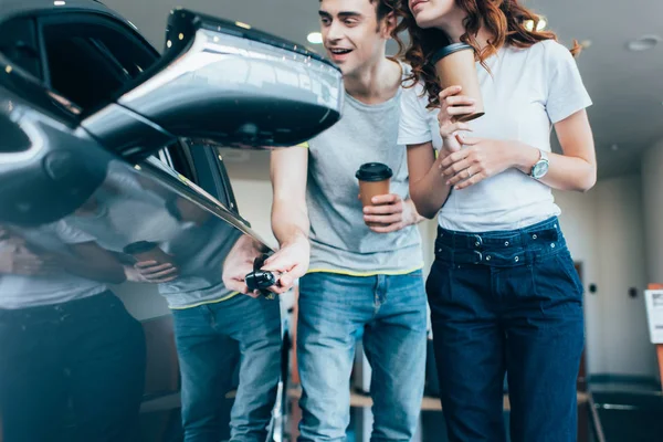 Обрізана дівчина, що стоїть біля чоловіка, тримає паперову чашку, дивлячись на автомобіль — стокове фото