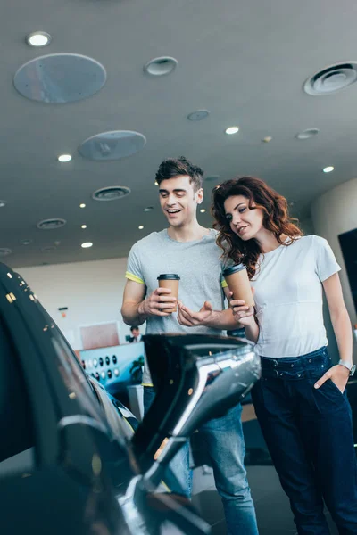 Cheerful man holding paper cup near curly woman standing with hand in pocket while looking at automobile — Stock Photo