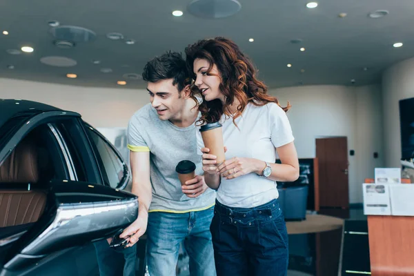 Cheerful man holding paper cup near curly girl while looking at automobile — Stock Photo