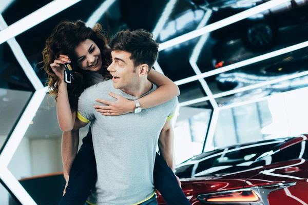 Handsome man piggybacking happy curly woman with keys in hand while standing near automobile — Stock Photo