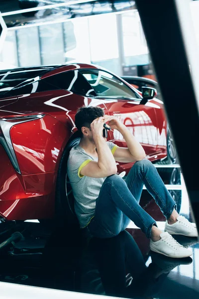 Upset man sitting near red automobile in car showroom — Stock Photo