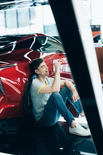 Messa a fuoco selettiva di uomo felice seduto e ridere vicino auto rossa — Foto stock