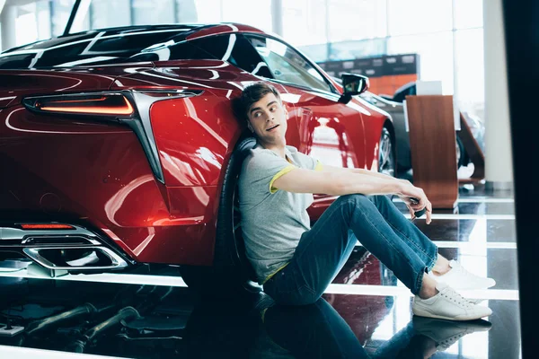 Handsome man sitting near red car in car showroom — Stock Photo