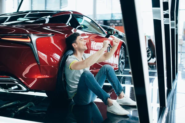 Foyer sélectif de l'homme heureux assis sur le sol près de la nouvelle voiture rouge et montrant geste oui — Photo de stock