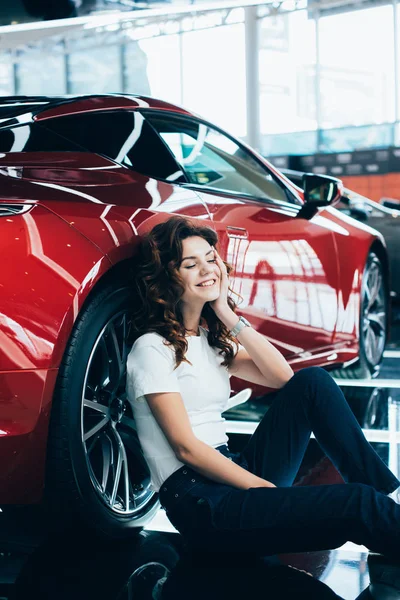 Selective focus of happy woman with closed eyes sitting on floor near new red car — Stock Photo