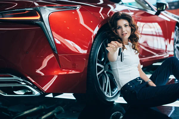 Щаслива жінка сидить на підлозі біля нової червоної машини і тримає ключі — стокове фото