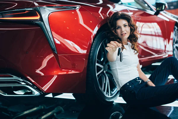 Mujer feliz sentado en el suelo cerca de nuevo coche rojo y la celebración de llaves - foto de stock