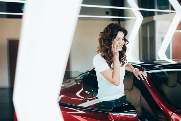 Selektiver Fokus einer lächelnden Frau, die auf dem Smartphone spricht, während sie in der Nähe eines neuen Autos steht — Stockfoto