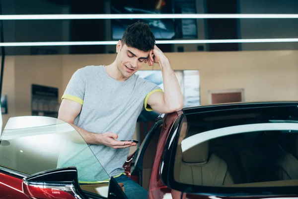 Селективный фокус улыбающегося мужчины с помощью смартфона, стоя рядом с машиной — стоковое фото