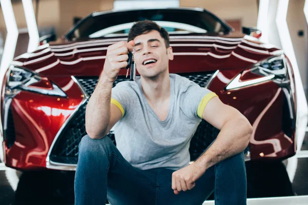 Selective focus of happy man sitting near new red car and holding car keys — Stock Photo