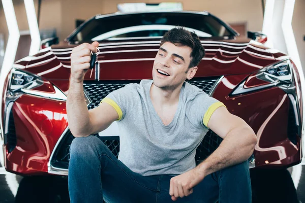 Selektiver Fokus des glücklichen Mannes, der Autoschlüssel hält, während er in der Nähe eines neuen roten Autos sitzt — Stockfoto