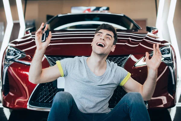 Feliz hombre emocionado sentado cerca de coche nuevo en sala de exposición de coches - foto de stock