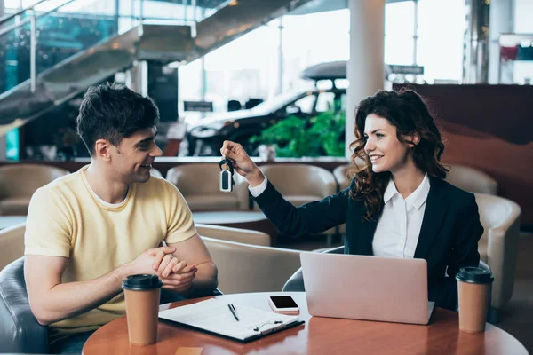 Lächelnder Autohändler gibt Autoschlüssel an glücklichen gutaussehenden Mann, während er am Tisch sitzt — Stockfoto