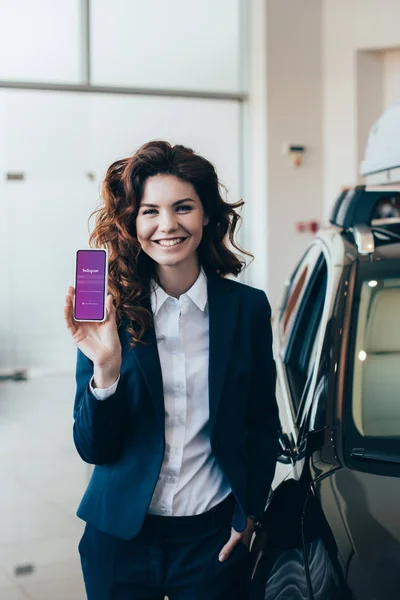 Smiling businesswoman holding smartphone with instagram app on screen and holding hand in pocket — Stock Photo