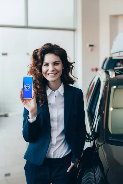 Smiling businesswoman showing smartphone with shazam app on screen and holding hand in pocket — Stock Photo