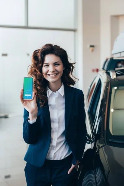 Attractive businesswoman holding smartphone with twitter app on screen and holding hand in pocket — Stock Photo