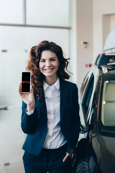 Femme d'affaires joyeuse tenant smartphone avec application netflix à l'écran et tenant la main dans la poche — Photo de stock