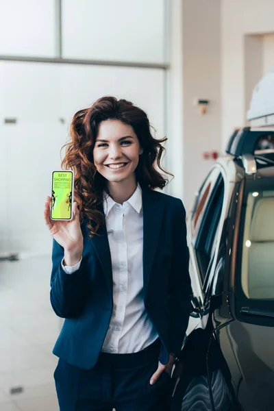 Bonita mujer de negocios sosteniendo el teléfono inteligente con la aplicación de compras en la pantalla y sosteniendo la mano en el bolsillo - foto de stock