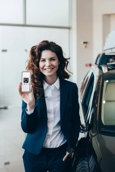 Alegre empresaria sosteniendo el teléfono inteligente con la aplicación uber en la pantalla y sosteniendo la mano en el bolsillo - foto de stock