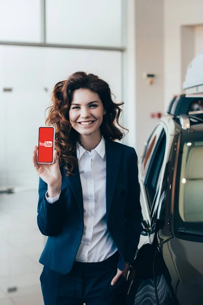Beautiful businesswoman holding smartphone with youtube app on screen and holding hand in pocket — Stock Photo