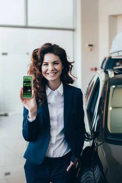 Smiling businesswoman holding smartphone with booking app on screen and holding hand in pocket — Stock Photo