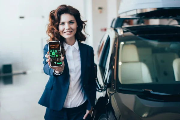 Selective focus of pretty businesswoman showing smartphone with graphs and charts on screen and holding hand in pocket — Stock Photo