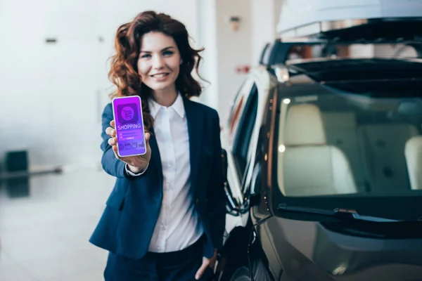 Избирательный фокус улыбающейся деловой женщины, держащей смартфон с приложением для покупок на экране и смотрящей в камеру — стоковое фото