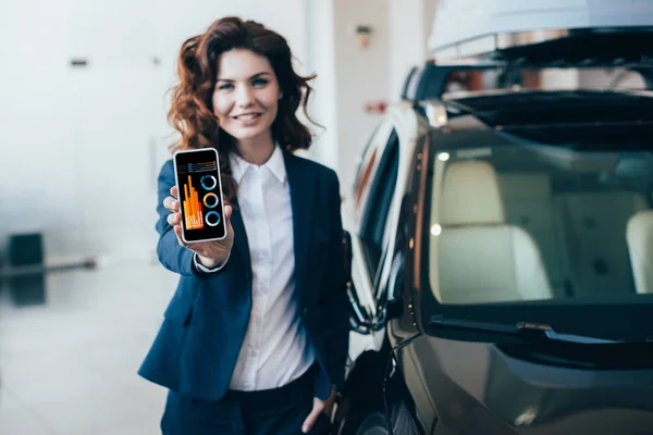 Selective focus of smiling businesswoman holding smartphone with graphs and charts on screen and looking at camera — Stock Photo