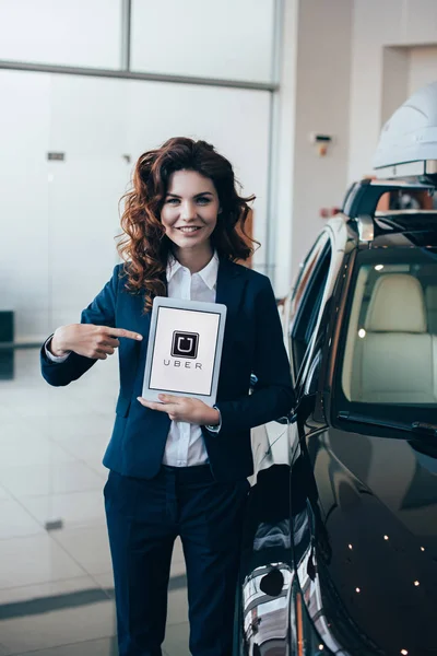 Mujer de negocios sonriente apuntando con el dedo a la tableta digital con la aplicación uber en la pantalla - foto de stock