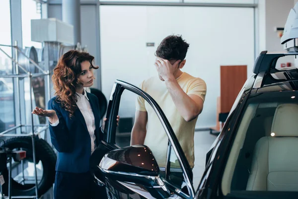 Pretty car dealer and man holding hand on face standing near new car — Stock Photo