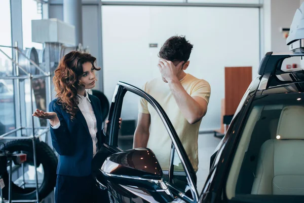 Joli concessionnaire de voiture et homme tenant la main sur le visage debout près de voiture neuve — Photo de stock